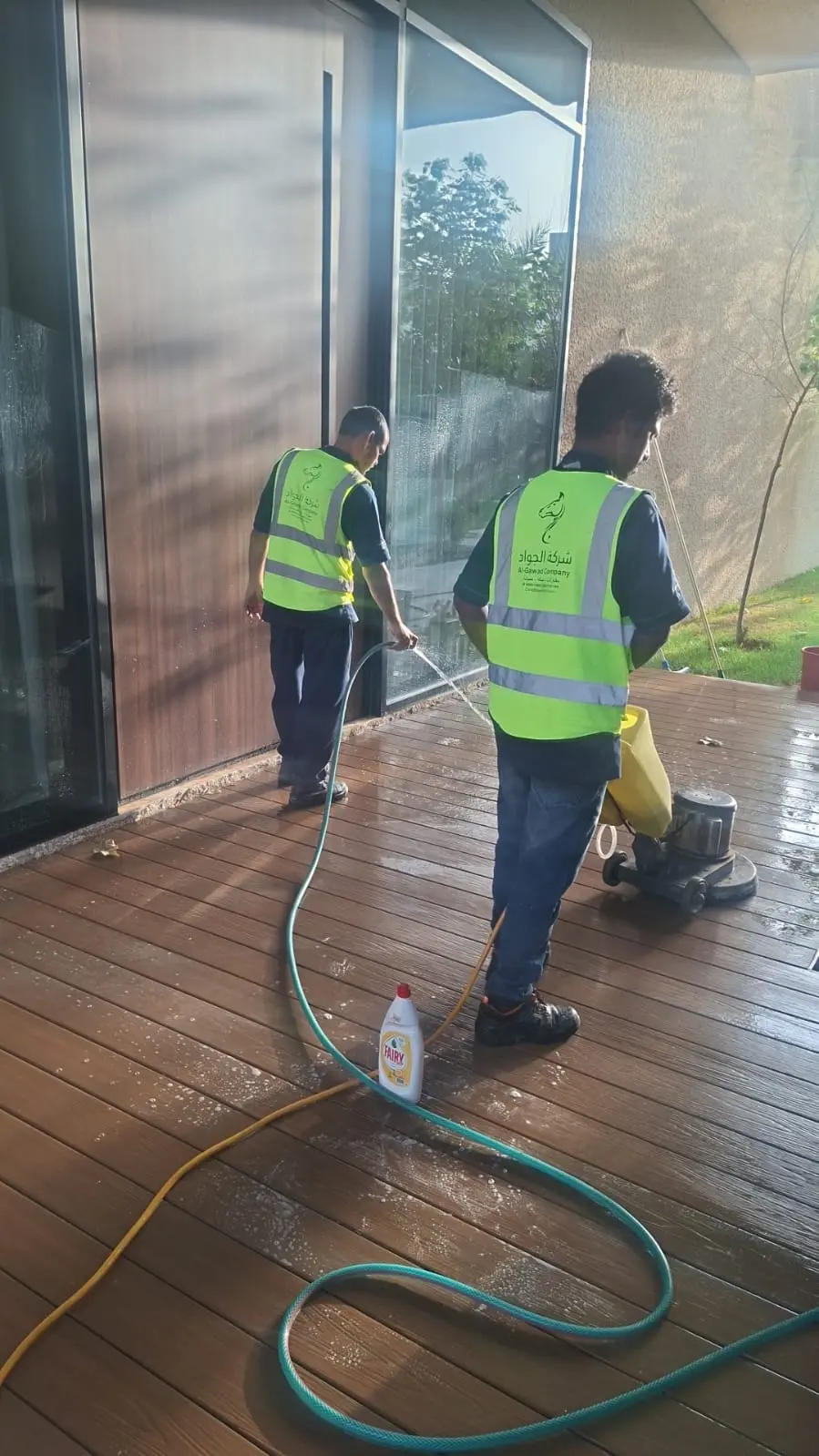 فني شركة الجواد يوجه مضخة مياه ذات ضغط عالي جداً (Pressure Washer) لتنظيف ممر من بلاط الإنترلوك المتداخل الأحمر والرمادي في حديقة فيلا بالرياض